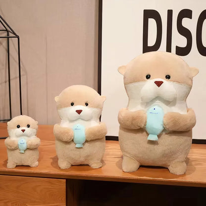 Three soft beige otter plushies of varying sizes holding a blue fish on a wooden table